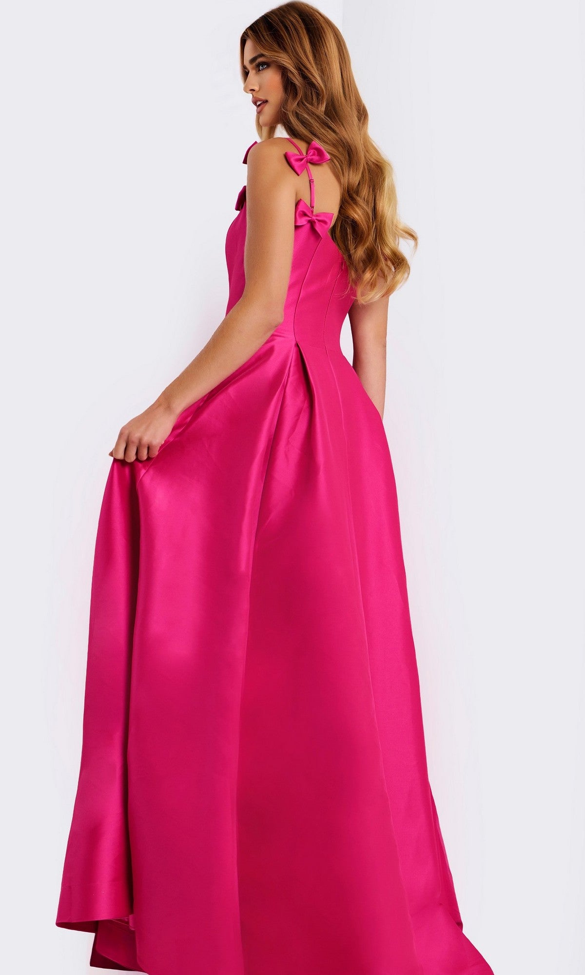 A woman stands holding the side of her Long Prom Dress: Jovani 46468, a bright pink satin gown with bow strap details and an elegant floor-length a-line skirt.